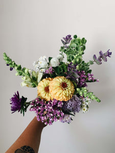 Miscellany + Co's Local Flower Bouquet subscription - a colorful bouquet with purples, whites, greens, and a hint of yellowy-orange toned zinnias held up against a beige wall