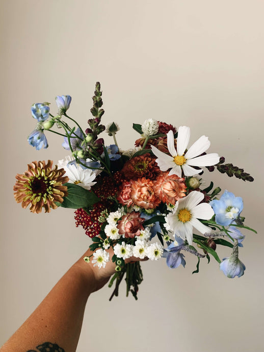 Miscellany + Co's Local Flower Bouquet subscription - a colorful bouquet with blues, whites, and peachy tones held up against a beige wall