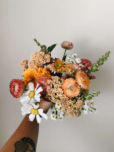 Miscellany + Co's Local Flower Bouquet subscription - a colorful bouquet with whites, peachy, coral, and orange tones held up against a beige wall