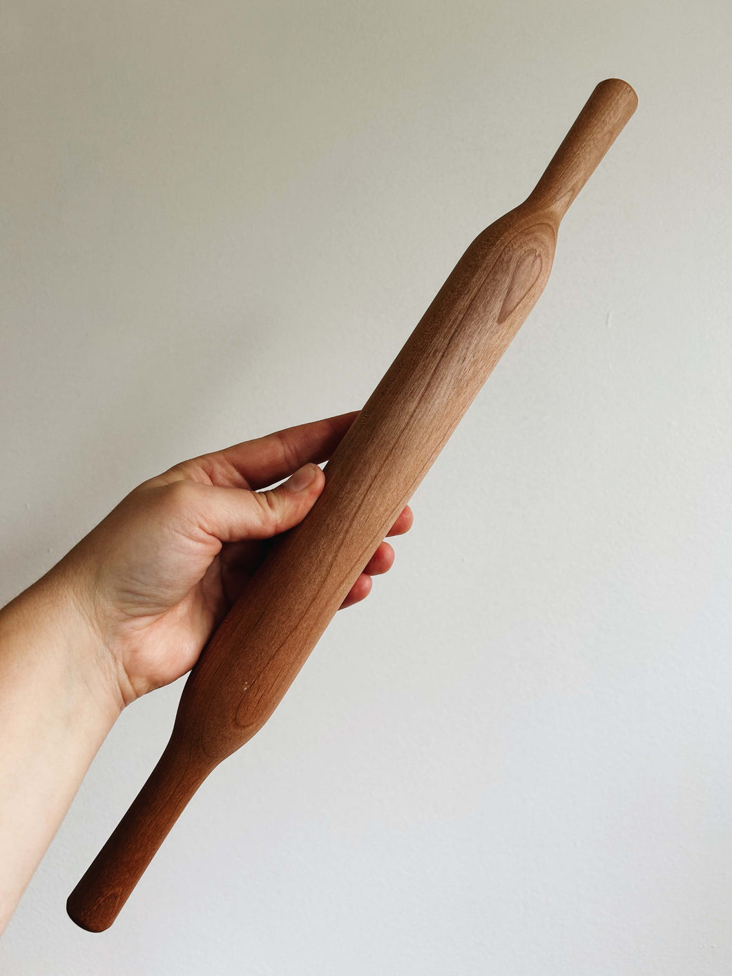 A hand holds a cherry wood rolling pin against a cream wall