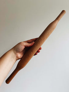 A hand holds a cherry wood rolling pin against a cream wall