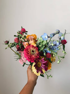 Miscellany + Co's Local Flower Bouquet subscription - a rainbow bouquet with blues, bright pinks, yellows, oranges, and green tones held up against a beige wall