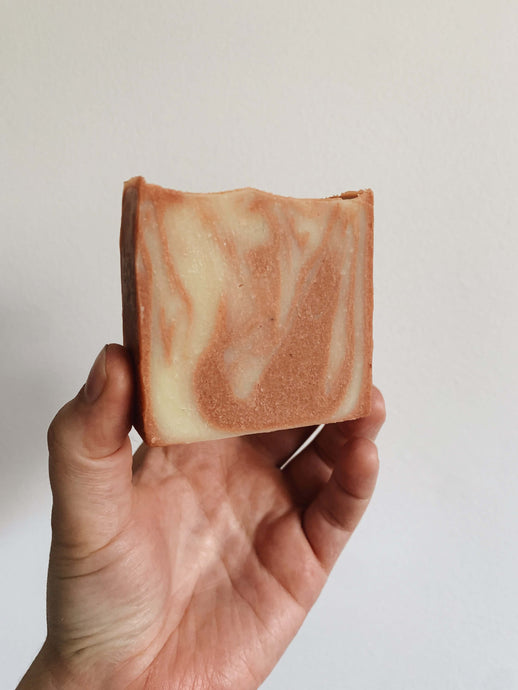 A peachy-pink and white swirled square soap bar held in Kitty's hand against a white wall