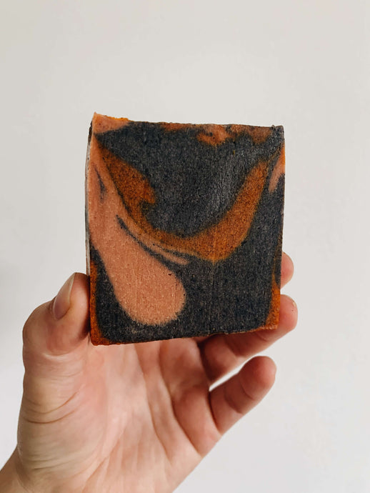 A peach, purple, and orange swirled square soap bar held up against a white wall -Tea Tree Grapefruit Soap Bar - Miscellany and Co