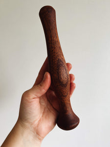 A hand holds a walnut wood cocktail muddler against a cream wall