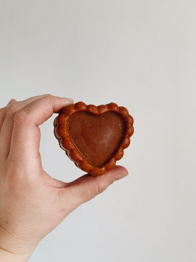 A brick red soap made in a heart shape being held by a hand against a white wall
