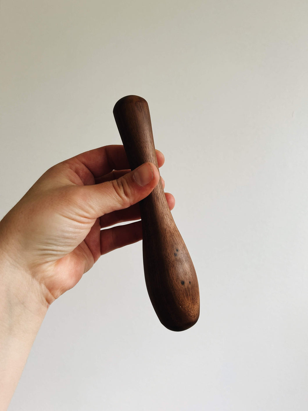 A hand holds a walnut wood cocktail muddler against a cream wall