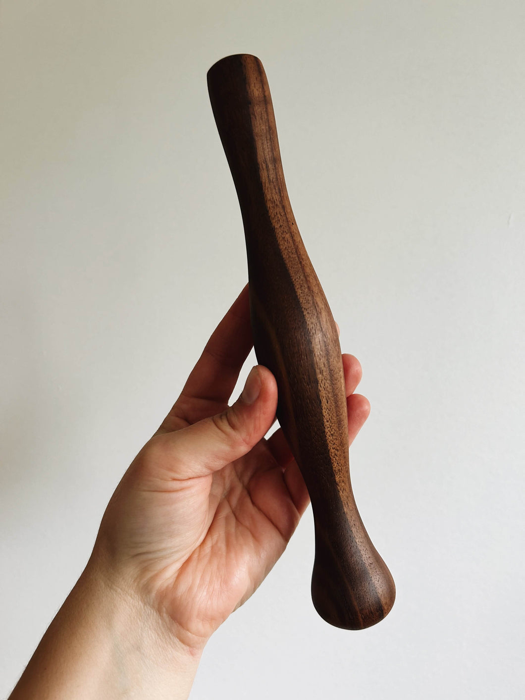 A hand holds a walnut wood cocktail muddler against a cream wall