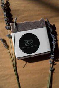 A picture of a purple and slightly speckled soap bar wrapped in a paper band and finished with a round black sticker with Miscellany and Co's house logo. There are sprigs of dried lavender resting beside both sides of the soap bar. The background is brown kraft paper. 