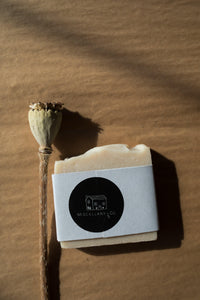 A picture of a cream soap bar wrapped in a paper band and finished with a round black sticker with Miscellany and Co's house logo. There is a dried poppy pod resting to the left of the soap bar. The background is brown kraft paper. 
