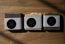 Load image into Gallery viewer, Three soap bars in a row - purple speckled bar, cream bar, and green bar, all wrapped in paper and and finished with a round black sticker with Miscellany and Co&#39;s house logo. Background is brown kraft paper.