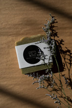 Load image into Gallery viewer, A picture of a green soap bar wrapped in a paper band and finished with a round black sticker with Miscellany and Co&#39;s house logo. There is a sprig of dried sea lavender resting beside the soap bar. The background is brown kraft paper. 