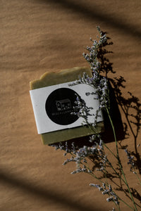 A picture of a green soap bar wrapped in a paper band and finished with a round black sticker with Miscellany and Co's house logo. There is a sprig of dried sea lavender resting beside the soap bar. The background is brown kraft paper. 